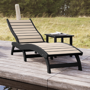 Black curved lounge chair and side table on a wooden deck with grass and trees in the background.