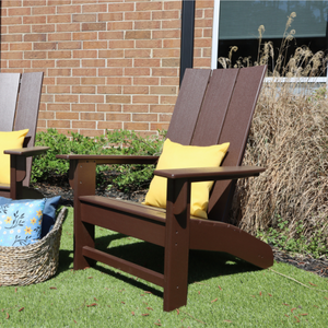 Brown Adirondack chair with yellow cushions on a grassy area next to a brick wall.